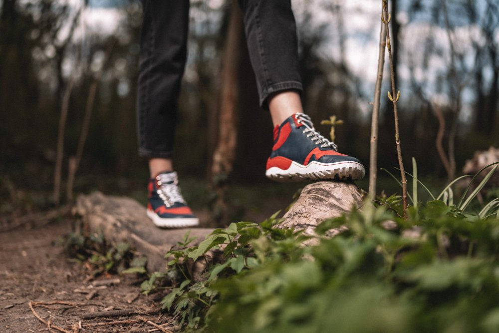 Osoba v čiernych nohaviciach a TREQ Red Waterproof teniskách vyrobených ako barefoot topánky od ZAQQ stojí v lese na prevrátenom kmeni stromu. Zelené rastliny obklopujú kmeň stromu, v rozmazanom pozadí sú viditeľné stromy.