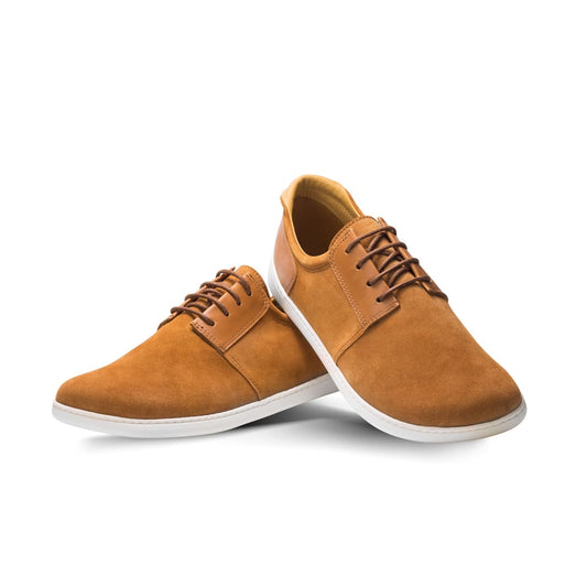 Ein Paar PIQUANT Sierra-Schuhe von ZAQQ aus braunem Premium-Veloursleder mit weißen Gummisohlen und dunkelbraunen Schnürsenkeln. Diese Barfuß-Sneaker sind kreativ positioniert, indem ein Schuh seitlich an den anderen gelehnt ist, vor einem makellos weißen Hintergrund.