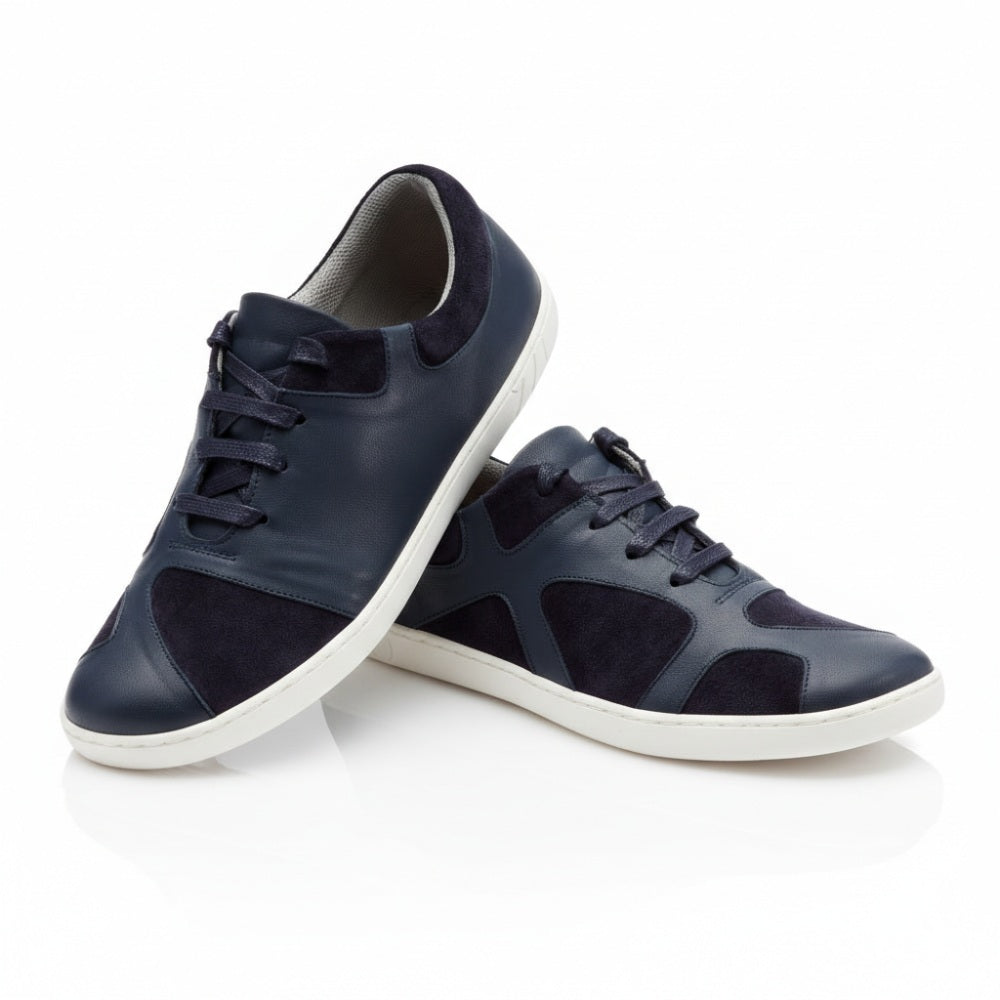 Der QAAANAAQ Blue von ZAQQ ist ein Paar stylischer marineblauer Sneaker mit weißer Sohle, gefertigt aus Leder und Veloursleder. Ein Schuh ist leicht auf dem anderen gestützt, um sein Design und seine Strukturdetails hervorzuheben.