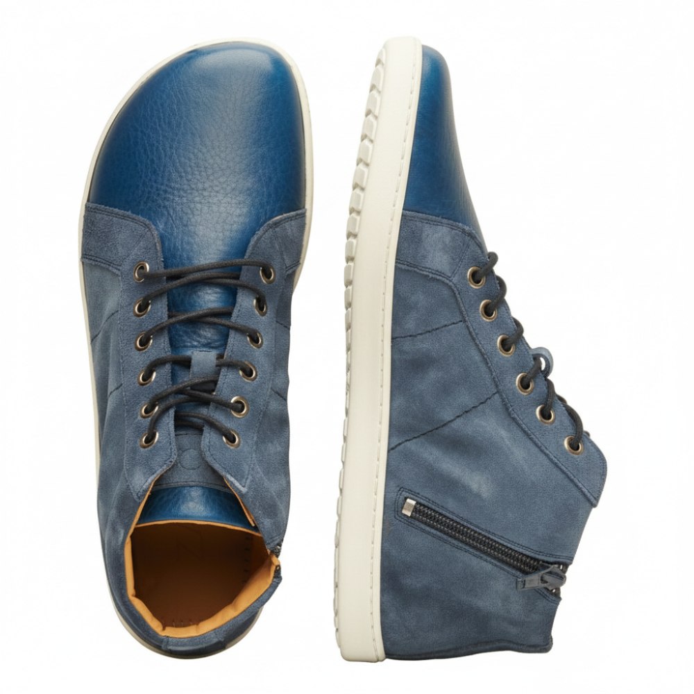 Zwei ZAQQ QORE Mid Deep Sea High-Top-Schuhe aus blauem Leder, wobei ein Schuh nach oben und der andere von der Seite zeigt. Die Schuhe verfügen über schwarze Schnürsenkel, die durch silberne Ösen und Veloursleder-Einsätze verflochten sind. Ein Reißverschluss ziert die Seite eines Schuhs, während die Sohle den Markennamen ZAQQ prominent zur Schau stellt.