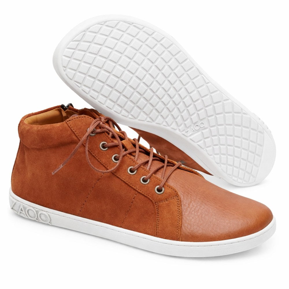 Der QORE Mid Sierra von ZAQQ strahlt mit seinem High-Top-Design aus braunem Leder und den wunderschön kontrastierenden weißen Sohlen Eleganz aus. Ein Schuh steht aufrecht, während der andere ein strukturiertes Muster auf der Sohle aufweist, das die verschiedenen Lederarten hervorhebt, die bei seiner Herstellung verwendet wurden.